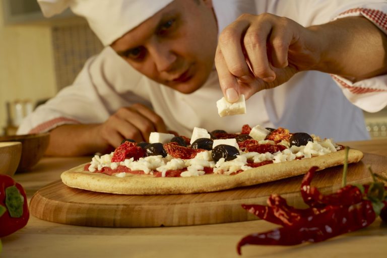 Chef adding seasoning to a pizza topped with tomatoes, olives, and cheese on a wooden board.