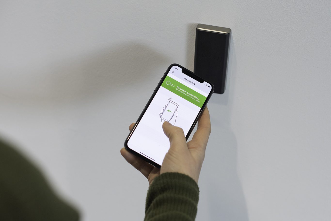 A person holds a smartphone near a wall-mounted device, displaying a verification screen.