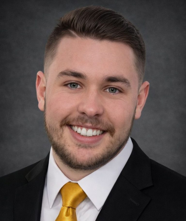 Smiling man in a suit with a yellow tie, against a grey background.