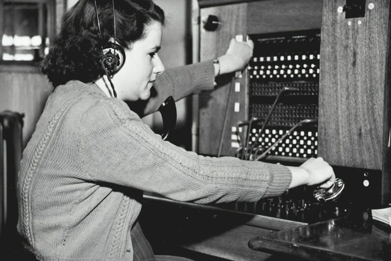 Smiling woman with a headset in an office, with a man working on a computer nearby.