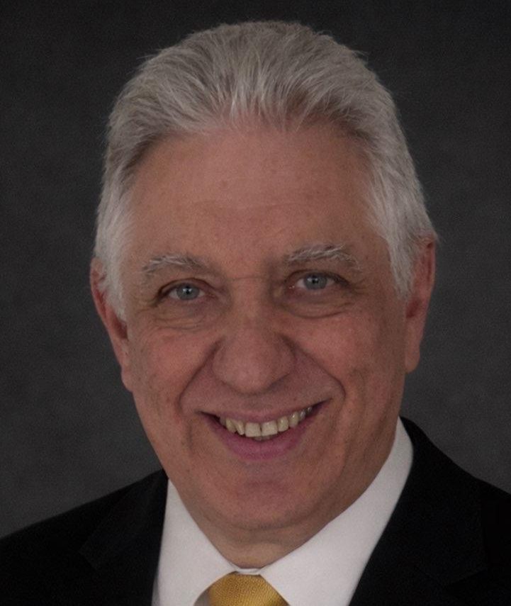 Smiling older man in a suit with a light tie against a dark background.